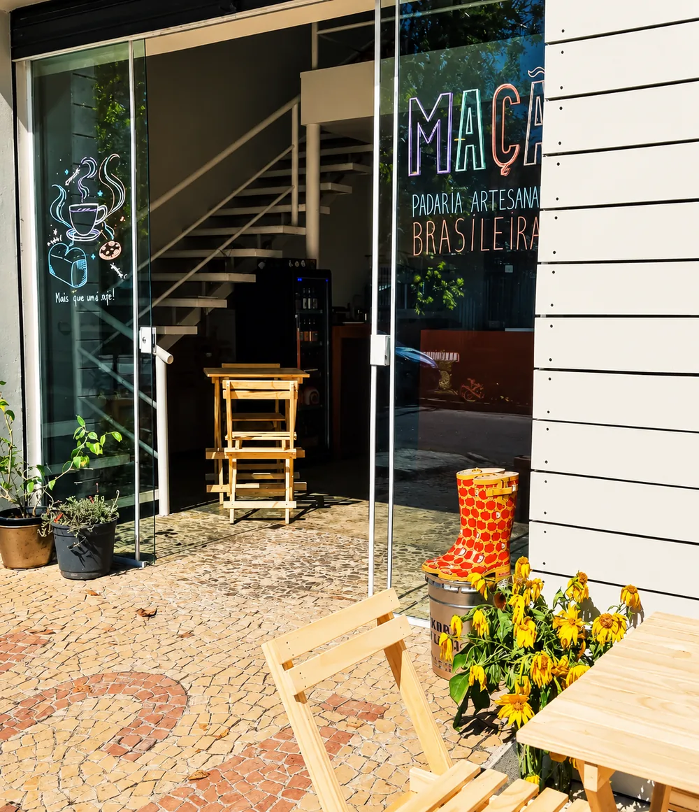 Fachada da Maçã com porta de vidro, escrito Maçã padaria artesanal brasileira, cadeiras de madeira e galochas com estampa de maçã