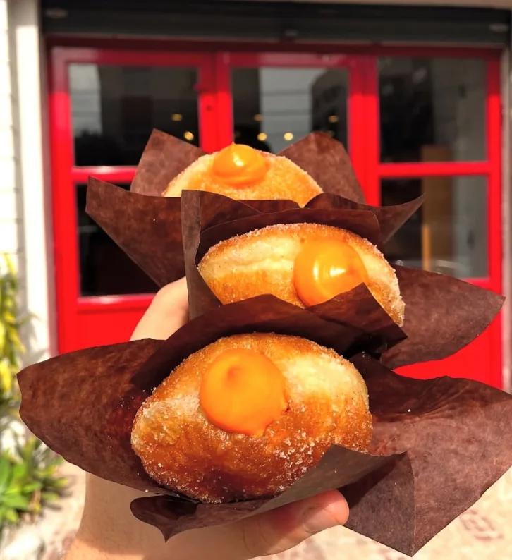 Três sonhos açucarados com creme laranja em papel marrom, em frente à porta vermelha da loja