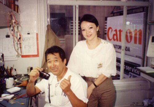 Jorge Namba sentado à bancada, segurando uma câmera, com Dona Carmen ao lado, numa foto antiga da oficina